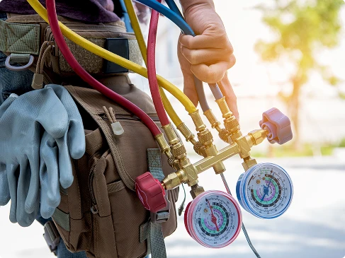 A person holding HVAC manifold gauges and hoses, with gloves and a tool bag visible, likely preparing for air conditioning maintenance or repair work.