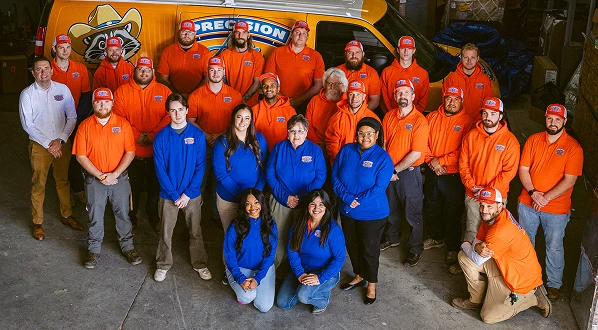 A group of 23 people pose indoors in front of a yellow van; most wear orange shirts and red hats, while five women in blue tops kneel or stand in the front row.