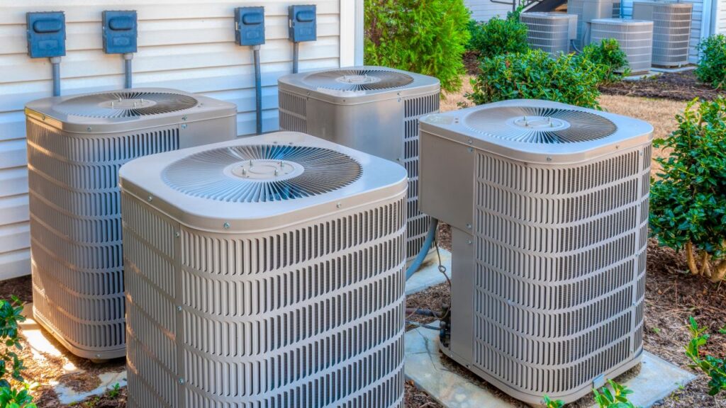 Four outdoor air conditioning units are installed beside a building, surrounded by shrubs and electrical boxes on the exterior wall. AC Unit Replacement Services